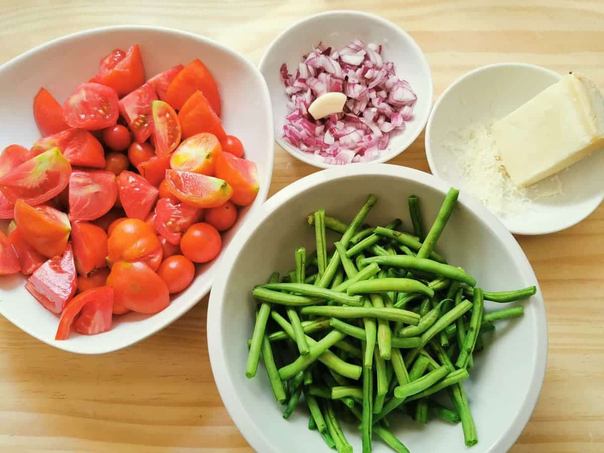 Sliced tomatoes in white bowl, blanched green beans in white bowl. Peeled and chopped onions and garlic in white bowl and grated cheese in white bowl.