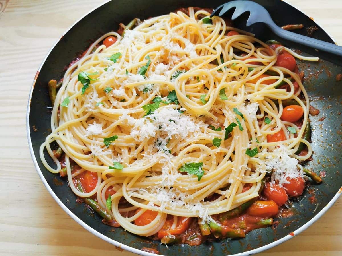 Cooked spaghetti with grated cheese and chopped parsley on top of green bean and tomato sauce in skillet.