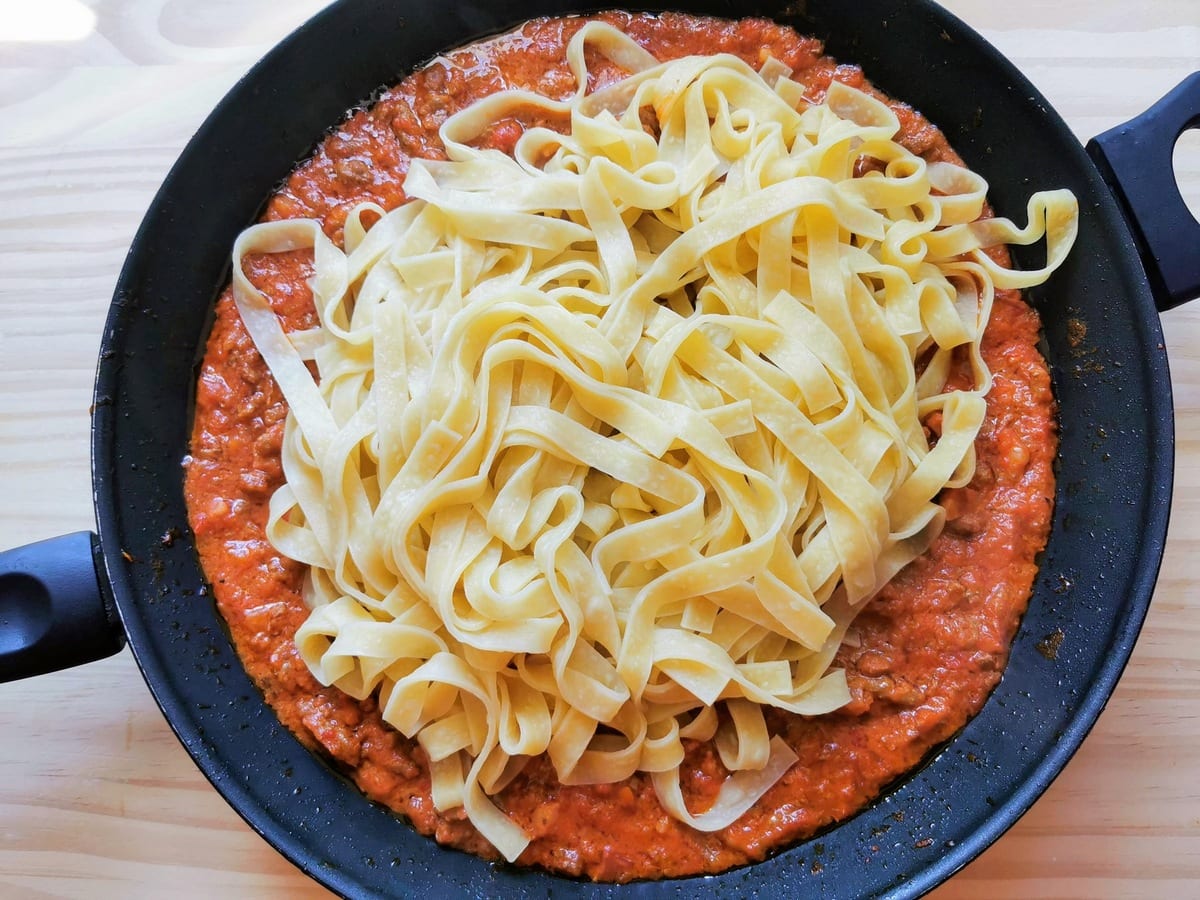 Freshly cooked tagliatelle added to the bolognese sauce in frying pan.