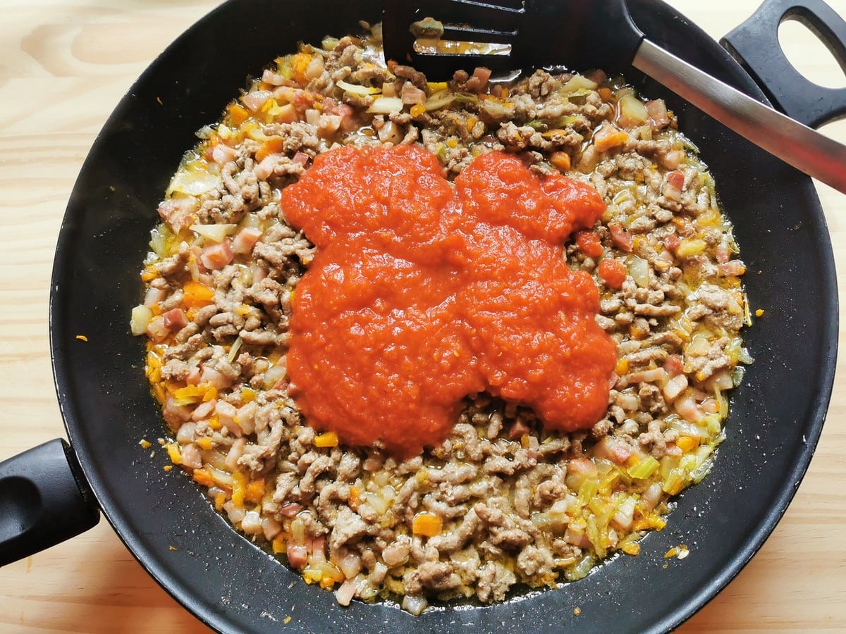 Adding stock and tomatoes to the ground meat and vegetables in frying pan.