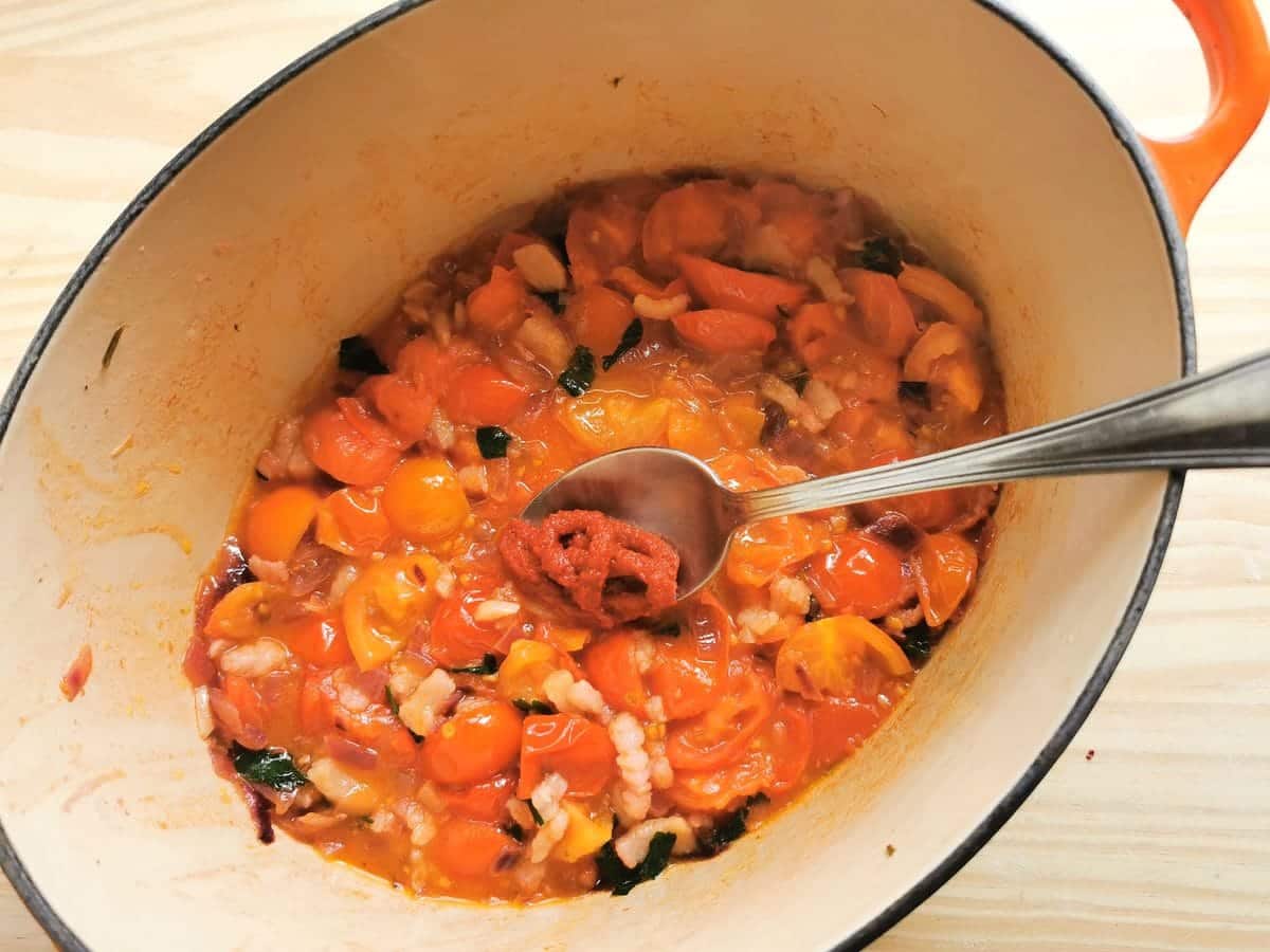 Tablespoon with tomato concentrate in Dutch oven with cooked tomatoes, onion and lardo